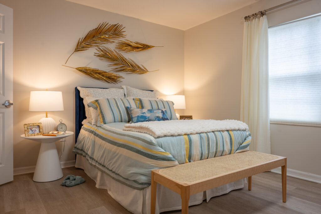 A cozy bedroom with striped bedding and palm frond wall art offers peaceful accommodations for senior living near Winter Park FL.